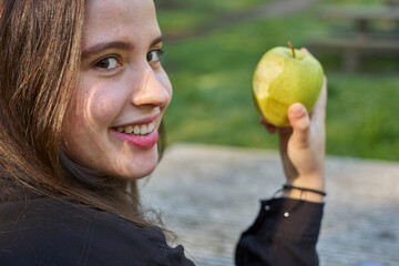 beautiful young woman in a black shirt sitting on a stone table eating an apple in nature with her mobile, a coffee and her bag with trees in the background. vegan teen taking a break in the park