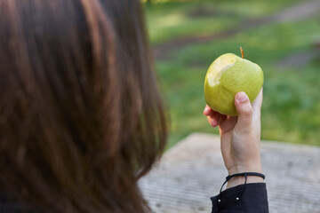 beautiful young woman in a black shirt sitting on a stone table eating an apple in nature with her mobile, a coffee and her bag with trees in the background. vegan teen taking a break in the park