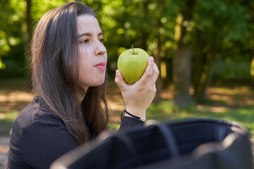 beautiful young woman in a black shirt sitting on a stone table eating an apple in nature with her mobile, a coffee and her bag with trees in the background. vegan teen taking a break in the park