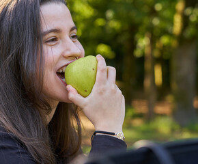 beautiful young woman in a black shirt sitting on a stone table eating an apple in nature with her mobile, a coffee and her bag with trees in the background. vegan teen taking a break in the park