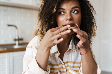 The black woman eating a toast with a chocolate paste and looking to the side while sitting in the kitchen