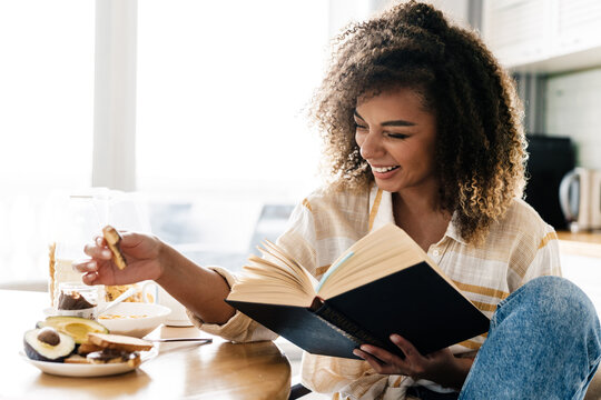 The Side View Of A Laughing Woman Holding A Book In Her Hands And Reaching For A Cookie While Sitting In The Kitchen