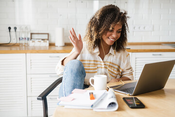 A smiling black woman in a wired headphones waving at the laptop camera while sitting at the table in the kitchen