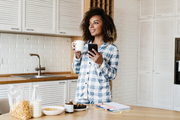 The smiling black woman in pajamas with a cup and a phone in her hands standing in the kitchen