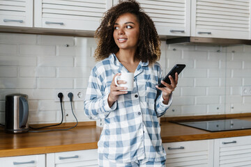 The portrait of a smiling black woman in pajamas looking to the side with a cup and a phone in her...