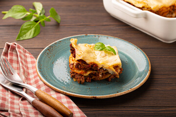 Slice of homemade traditional Italian lasagna with beef bolognese ragout and cheese served on blue ceramic plate on dark brown rustic wooden table, angle view 