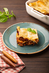 Slice of homemade traditional Italian lasagna with beef bolognese ragout and cheese served on blue ceramic plate on dark brown rustic wooden table, angle view 