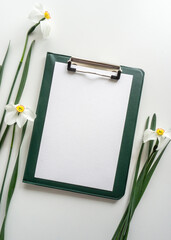 Green clipboard with daffodils on white background, home office workspace flat lay.