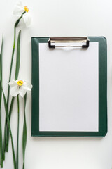 Green clipboard with daffodils on white background, home office workspace flat lay.