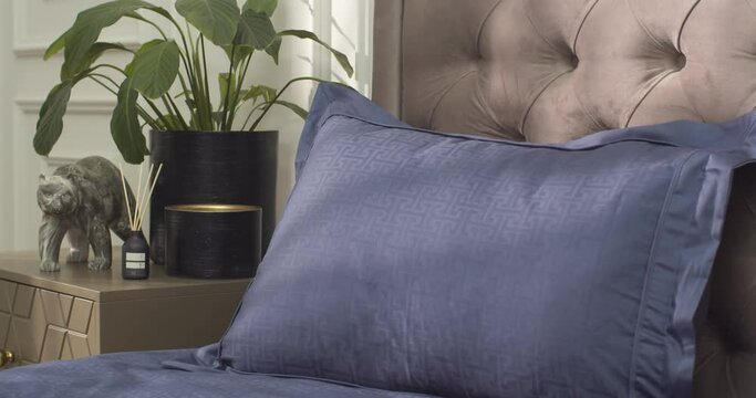Side close-up of two dark blue pillows with floral pattern and blue cover on the large double bed padded with brown soft material. There is a blurred bedside table with flower in the pot and bear stat