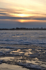 A landscape view of the frozen sea during ice drift in spring with dramatic sky and with reflections in melt water in the evening at sunset. In the distance, the black strip of the island. © Helena