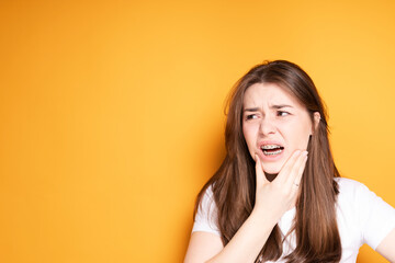 Sad girl in braces touches her jaw suffering from pain in her teeth while standing on a yellow...