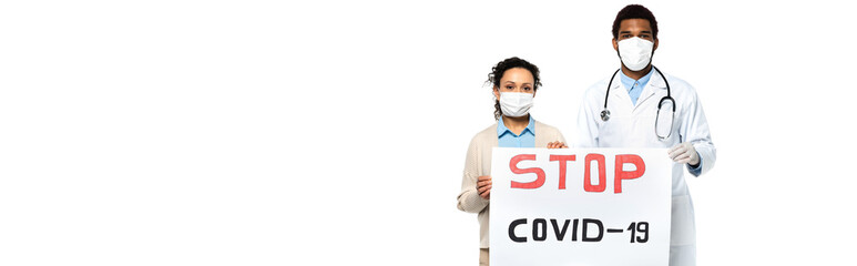 African american patient and doctor in medical masks holding placard with stop covid-2019 lettering isolated on white, banner