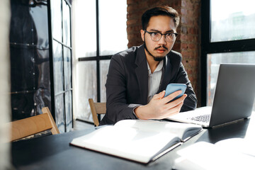 Serious korean businessman in glasses sits at a table with a laptop and documents and holds a phone in his hand