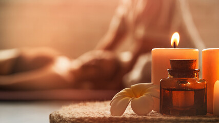 Warm inviting picture of beautiful spa composition with frangipani flower, oil flask, and candles. Blurred figures of the masseur and his guest in background.