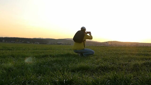 Young caucasian man in yellow jacket with backpack taking photo with mobile phone at sunset in czech spring landscape