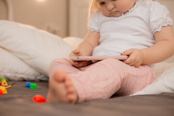 Baby blonde girl plays with the phone at home on the bed. Children and technology