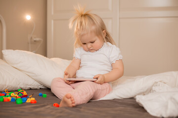 Baby blonde girl plays with the phone at home on the bed. Children and technology