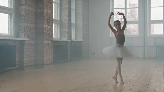 Full shot of beautiful and talented 9-year-old ballerina in tutu and pointes dancing graciously in smoke-filled choreography class