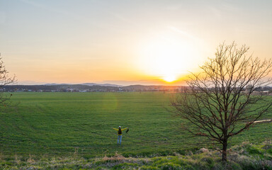 Obraz na płótnie Canvas Aeirial view young caucasian man in yellow jacket with backpack standing on meadow, field at sunset in czech spring landscape