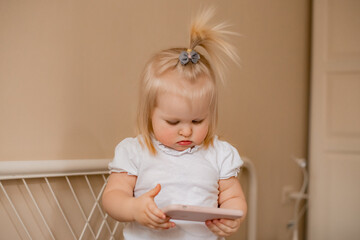 Fototapeta premium Baby blonde girl plays with the phone at home on the bed. Children and technology