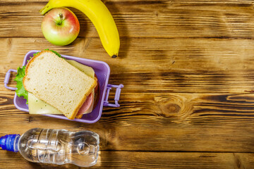 Lunch box with sandwiches, bottle of water, banana and apple on a wooden table. Top view, copy space