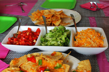 Close-up of a table set for a party, with nachos, a dish of taco toppings, a pink table cloth and napkins, place mats and paper plates.