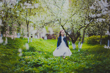 Little girl in black jacket and long white dress running in spring cherry garden. Happy child among white flowers trees. Childhood. Young lady walking in sunny blooming park at springtime.