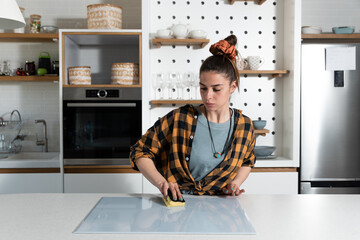Young single housewife washes and cleans the cook top hob from a modern stove in with a sponge...