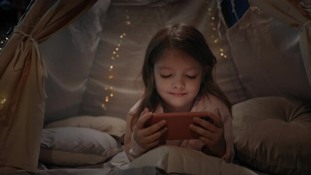 Cheerful little girl spending time in decorative tent with lightings in evening playroom. Kid lying on floor and smiling while watching cartoon on smartphone. Concept of childhood.
