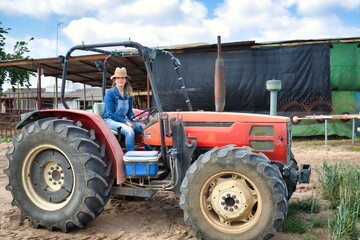 Blonde woman wearing a hat and jeans riding on a red tractor ready to work in the field