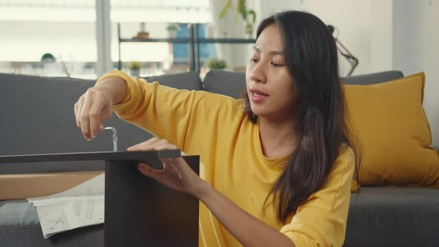 Happy asian young woman unpacking box and reading the instructions to assemble new furniture decorate house build table with carton box in living room at home.