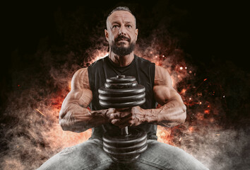 Muscular man posing with a dumbbell on a background of fire and smoke. Fitness and bodybuilding concept.