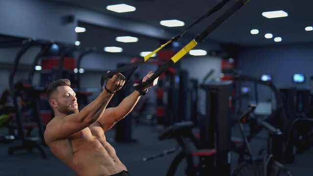 Sportive guy in the gym. Muscular man without shirt pumping muscles on a simulator with a rubber at sports centre. Daily workout of bodybuilder.