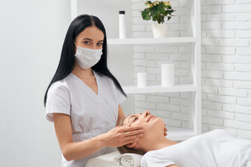 Side view of young brunette beautician in protective mask doing massage face for smiling young...