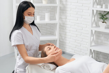 Obraz premium Side view of young brunette beautiful woman on procedure massage for face in beautician. Concept of special care for face and body in professional spa salon. 