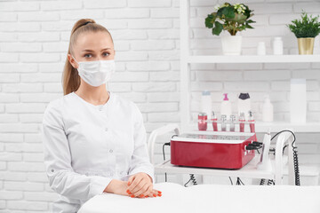 Front view of young beautiful beautician in white uniform and protective mask sitting and waiting...