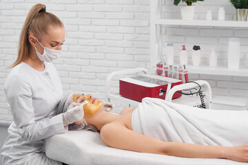 Obraz na płótnie Canvas Side view of young beautician in protective mask and rubber gloves doing special cosmetic mask for young beautiful patient. Concept of procedure for improvements skin in professional salon.