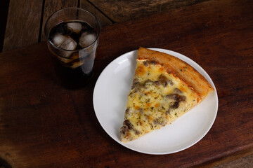 Top view of one slice or piece of pizza of meat or rump or filet, on a white plate, on a wooden table, next to a glass of soda or coke