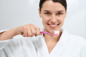 Close up portrait of happy young cute brunette woman in white robe holding toothbrush on light background. Concept of preparing for cleaning teeth with special paste at home. 