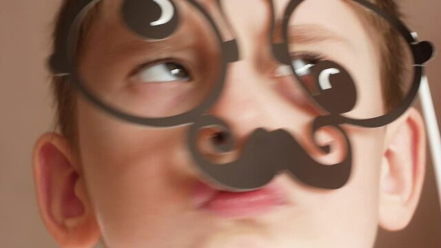 A cheerful, funny preschooler holds a mustache and glasses in front of his face, the child portrays a strict parent. A child's joke about an adult. Father's day humor. Selective focus,