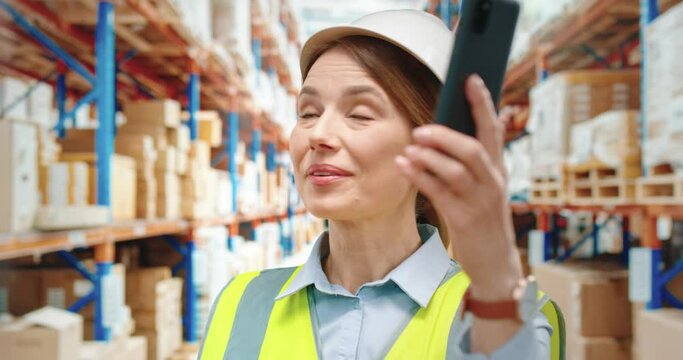 Beautiful female warehouse worker in white helmet talks on video call and shows large rows with cargo using smartphone .
