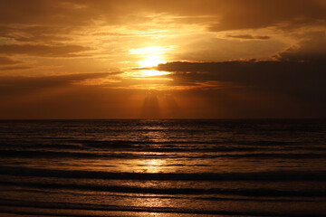 Fototapeta premium Sunset on a beach, orange sun is shining through the clouds and reflected in dark waves. Evening sea background for romantic travel