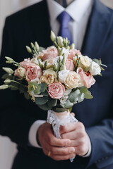 Young groom in blue blazer with blue tie holding bouquet with pink and biege roses and green...