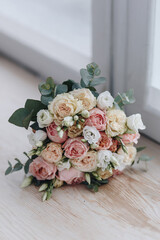 Brides bouquet made from white, pink and biege roses and eucaliptus branches laying on the wooden table near the window, close up vertical photo