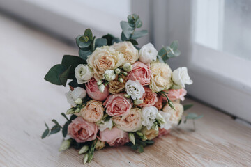 Brides bouquet made from white, pink and biege roses and eucaliptus branches laying on the wooden table near the window, close up photo