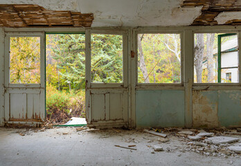 Abandoned house interior. Gallery with door and window frames, broken glass and fallen plaster, overgrown autumn garden seen through the windows