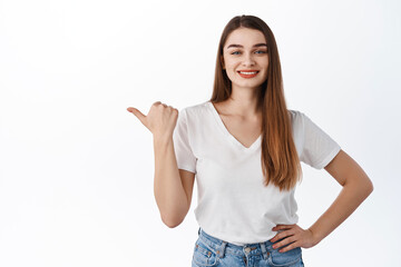 Smiling cheerful woman points thumb left, shows copy space advertisement, store promo deal, look determined, recommend product, stands against white background