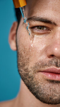 Half Face Closeup Of Attractive Man With Smooth Skin Looking At Camera, Applying Serum Beauty Product On His Face Using Glass Pipette Isolated On Blue Background