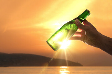 Hand raising a bottle of fresh beer backlit at sunset